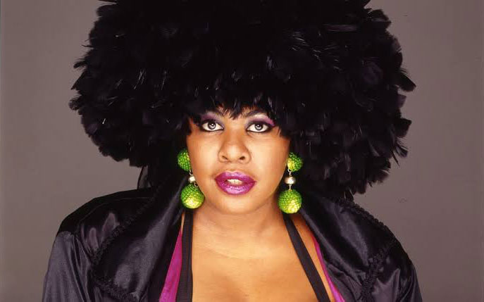Imagem de uma mulher negra olhando para frente com o chapéu de penas na cabeça e brincos redondos verdes.