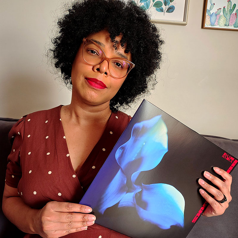 Imagem de uma mulher negra olhando de frente e segurando a capa de um disco de vinil da banda Bambara
